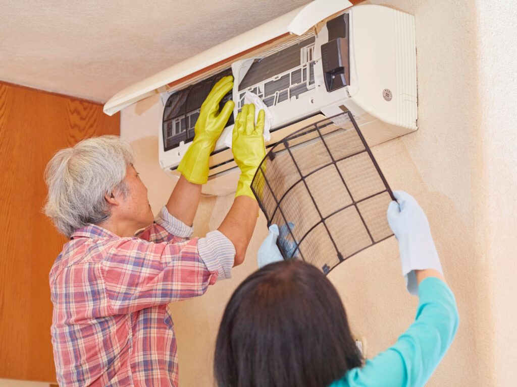 Two people cleaning a wall-mounted AC unit and removing the air filter.
